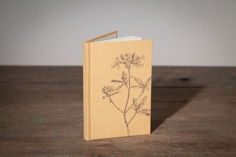 Op de afbeelding: Een klein, tan linnen notitieboekje met een bloemenprint op de voorkant. Het ontwerp toont een enkele stengel met bladeren en bloemen.