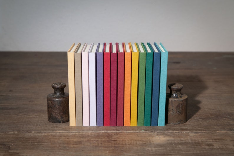 Op de afbeelding: Een rij van 14 kleurrijke hardcover notitieboekjes met linnen omslagen, gerangschikt op een houten oppervlak. De notitieboekjes zijn in verschillende tinten beige, roze, paars, rood, bruinrood, oranje, geel, groen, blauw en turquoise.