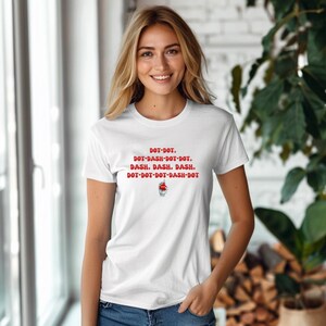 May include: White t-shirt with red text: DOT-DOT, DOT-DASH-DOT-DOT, DASH, DASH, DASH, DOT-DOT-DOT-DASH-DOT. The shirt features a small graphic below the text. The model is smiling.