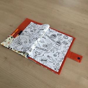 Könnte beinhalten: Ein weißes Stoffbuch mit einem schwarz-weißen Superhelden-Thema. Das Buch ist geöffnet, um eine leere Seite zum Ausmalen zu zeigen. Das Buch hat einen roten Stoffbezug und einen blauen Knopf.