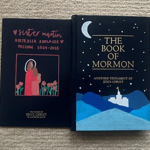 Könnte beinhalten: Zwei schwarze Bücher. Eines trägt den Text "sister martin AUSTRALIA ADELAIDE MISSION 2024-2025" mit einem Herz und einer Illustration. Das andere Buch trägt den Titel "THE BOOK OF MORMON" mit Mond, Sternen und einer Landschaft.