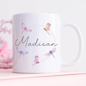 Pode incluir: Caneca de cerâmica branca com um círculo dourado e um desenho de libélulas em aquarela. O nome "Madison" está escrito em itálico preto dentro do círculo.