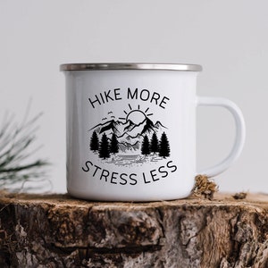 Puede incluir: Taza de camping de esmalte blanco con borde plateado. La taza presenta el texto "Hike More, Stress Less" en negro, junto con un gráfico de montaña y árboles. El diseño está impreso en ambas caras. La taza está sobre una superficie de madera.
