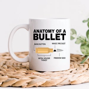 Pode incluir: Caneca de cerâmica branca com um gráfico preto de uma bala e o texto "Anatomia de uma bala". O gráfico mostra a bala com rótulos para "Botão Bang", "Coisa de suporte metálico", "Poeira de fogo mágica" e "Semente de liberdade".