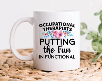 Pode incluir: Caneca de café de cerâmica branca com um design preto que diz "Occupational Therapists Putting the Fun in Functional". O design inclui uma ilustração floral em rosa, azul e verde.
