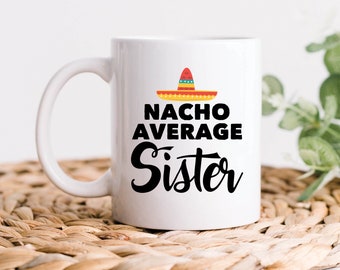 May include: White ceramic mug with a black and white design that says "Nacho Average Sister" with a colorful sombrero on top. The mug is sitting on a woven placemat.
