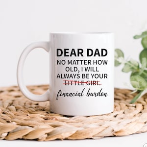 May include: White ceramic coffee mug with black text that reads "Dear Dad No matter how old, I will always be your little girl financial burden". The word "little" is crossed out and the word "financial" is underlined. The mug is sitting on a woven straw mat.