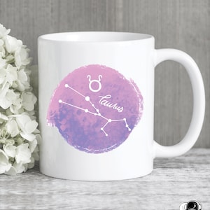 May include: White ceramic mug with a watercolor design of the Taurus constellation in pink and purple. The constellation is outlined in white and the word "Taurus" is written in white cursive below the constellation.