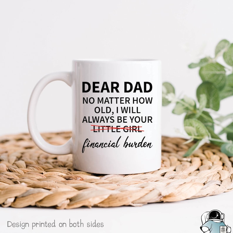 May include: White ceramic coffee mug with black text that reads "Dear Dad No matter how old, I will always be your little girl financial burden". The word "little" is crossed out and the word "financial" is underlined. The mug is sitting on a woven straw mat.