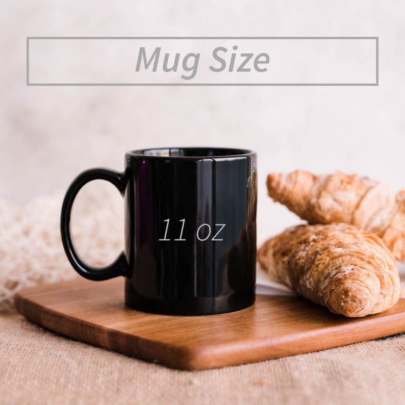 May include: A black ceramic mug with a white inscription that reads "11 oz". The mug is sitting on a wooden cutting board with two croissants.