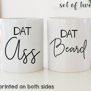 May include: Two white ceramic mugs with black text. One mug says "DAT Ass" and the other says "DAT Beard" in a handwritten font. The phrase "set of two mugs" is above the mugs. The phrase "Design printed on both sides" is below the mugs.
