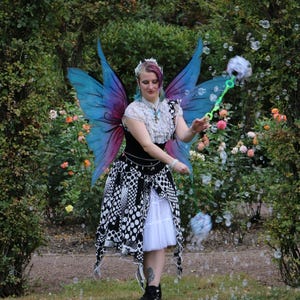 May include: A person in a fairy costume with blue and purple wings, a black and white patterned skirt, and a white top, blowing bubbles in a garden setting. The background features green foliage and colorful flowers.