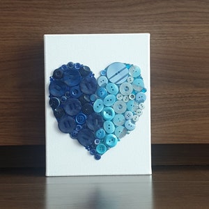 May include: A white canvas with a heart shape made of blue and light blue buttons.