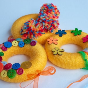 May include: Three yellow yarn wreaths decorated with colorful buttons, flowers, and pom-poms. The wreaths are tied with colorful ribbons.