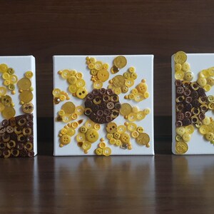 以下が含まれることがあります： 黄色と茶色のボタンでできたひまわり模様の小さな白いキャンバスが3枚です。