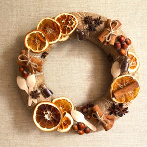 May include: A brown burlap wreath decorated with dried orange slices, cinnamon sticks, star anise, and small glass bottles. The wreath is accented with wooden spoons and hazelnuts.
