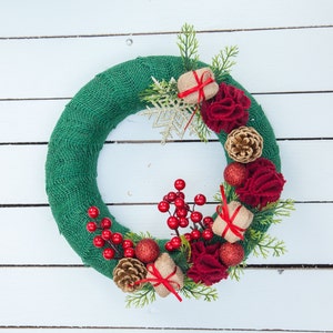 May include: A green burlap wreath decorated with red berries, red and white flowers, small wrapped gifts, and a gold snowflake.
