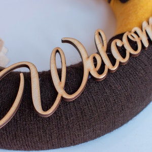 May include: A brown and yellow wreath with a wooden sign that says "Welcome" in a cursive font.