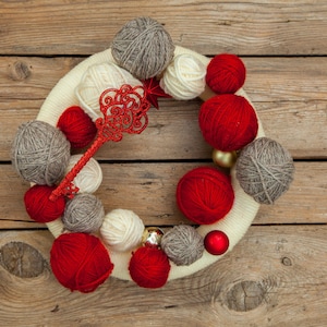 May include: A white wreath decorated with balls of yarn in red, white, and gray. The wreath is adorned with gold ornaments and a red sparkly key.