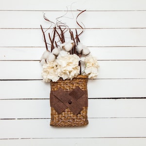 May include: A white wicker basket with a brown ribbon and a bouquet of white cotton and white flowers. The basket is hanging on a white wooden wall.
