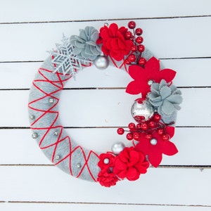 May include: A gray yarn wreath decorated with red felt flowers, silver glitter snowflakes, and silver ornaments.
