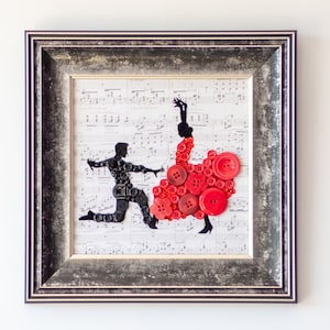以下が含まれることがあります： ボタンで作られた踊るカップルのシルエットが特徴の額装アート。男性の姿は黒、女性のドレスは赤です。背景は楽譜で、フレームはシルバーです。