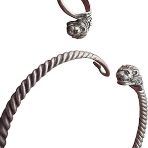 Puede incluir: Un conjunto de anillo y pulsera plateados. El anillo presenta un diseño de cabeza de león. La pulsera tiene un diseño de cuerda trenzada y terminales de cabeza de león. Las joyas se muestran sobre un fondo blanco.