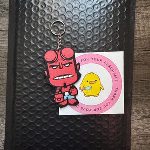 Op de afbeelding: Een rode Hellboy-sleutelhanger met een zilveren sleutelring wordt getoond op een zwarte luchtkussenenvelop. Een ronde sticker met een gele cartoonkuiken en de tekst "THANK YOU FOR YOUR PURCHASE!" is ook zichtbaar.