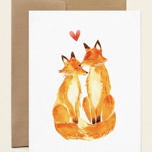 May include: Watercolor illustration of two orange foxes in a loving embrace, with a red heart above them. The foxes are sitting close together, with their heads touching. The artwork is on a white card, accompanied by a brown envelope.