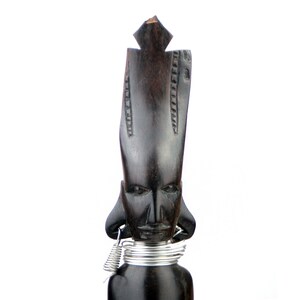 May include: A dark wood carved figurine of a person with a tall, pointed head and a silver wire necklace. The figurine has a simple, stylized face with large eyes and a small mouth.