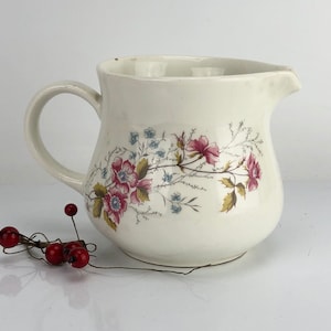 Puede incluir: Jarra de cerámica blanca con un diseño floral con flores rosas y azules. La jarra tiene un asa y un cuerpo redondeado.