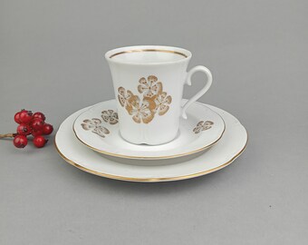 Könnte beinhalten: Ein weißes Porzellan-Kaffeetassen-Set mit goldenem Blumendesign. Die Tasse hat einen Henkel und die Untertasse einen erhöhten Rand.