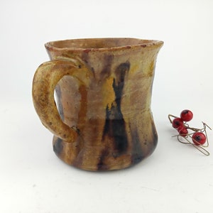 Puede incluir: Una taza de cerámica rústica de color marrón con asa. La taza tiene un esmalte jaspeado con vetas negras y amarillas.