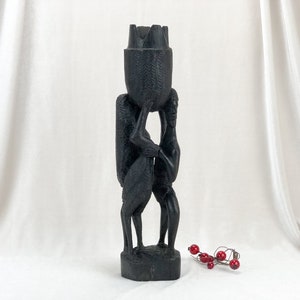 May include: A dark, carved wooden sculpture depicting two figures supporting a large, textured basket-like object. The sculpture is on a rectangular base and has a matte finish. A small string of red berries is in the foreground.