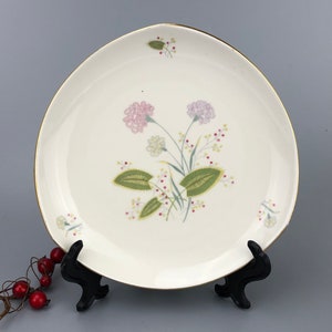 May include: A white ceramic plate with a gold rim features a floral design with pink and green flowers. The plate is on a black stand.