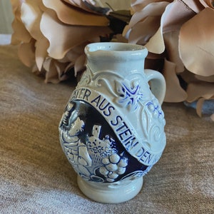 May include: A small, white ceramic pitcher with blue and white floral designs. The pitcher has a handle and a spout. The text "WATER AUS STEIN DEN" is written on the side of the pitcher.