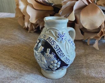 Puede incluir: Una pequeña jarra de cerámica blanca con diseños florales azules y blancos. La jarra tiene un asa y un pico. El texto "WATER AUS STEIN DEN" está escrito en el lateral de la jarra.