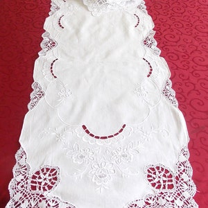 Könnte beinhalten: Weißer bestickter Tischläufer mit zarter Spitzenbordüre und Blumendesigns. Der Läufer liegt auf einer roten Tischdecke.