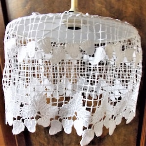 May include: White crocheted lampshade with a delicate floral design. The shade has a scalloped edge and is made of a fine, open weave.