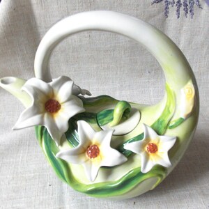 Könnte beinhalten: Eine weiße Teekanne aus Keramik in Form einer Blume mit grünen Blättern und weißen Blüten. Die Teekanne hat einen Henkel, der wie ein Schwanenhals geformt ist.