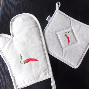May include: A set of two white linen pot holders with red chili pepper embroidery. The pot holder on the left is a mitten style and the pot holder on the right is a square shape.
