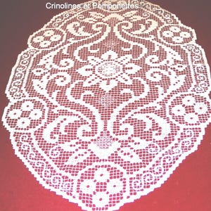 Könnte beinhalten: Ovale Tischdecke aus weißer Spitze mit aufwendigen Blumen- und Scrollmustern. Die Tischdecke hat ein filigranes, offenes Design mit einem gewellten Rand. Der Text "les collections de Crinolines et Pomponettes" befindet sich oben.