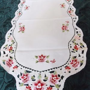 Può includere: Tovaglia da tavolo bianca con bordo a festoni e fiori rosa ricamati. Il testo "les collections de crinolines et pomponettes" è visibile nell'angolo in alto a sinistra.