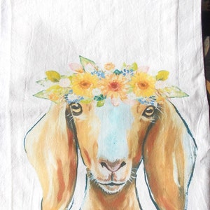 May include: White kitchen towel featuring a watercolor painting of a goat's face. The goat wears a floral crown of yellow sunflowers, blue flowers, and green leaves. The goat has brown fur and large ears. The artist's signature is in the bottom right corner.