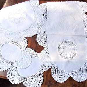 Puede incluir: Una colección de manteles individuales de encaje blanco vintage y un camino de mesa. Los manteles individuales tienen un diseño circular con un borde de ganchillo. El camino de mesa tiene un borde festoneado y un diseño central de encaje circular.