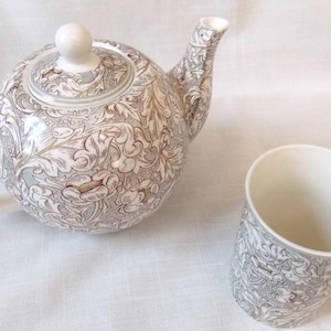 Puede incluir: Una tetera y una taza blancas con un patrón floral marrón. La tetera tiene una tapa y un pico. La taza tiene un asa.