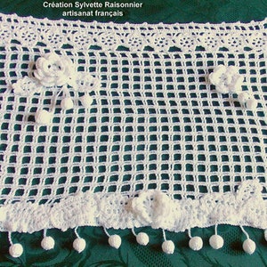 May include: White crocheted table runner with a delicate floral design and pom-pom trim. The runner is on a green tablecloth. "Creation Sylvette Raisonnier artisanat francais" is written on the runner.
