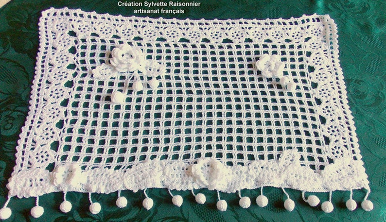 May include: White crocheted table runner with a delicate floral design and pom-pom trim. The runner is on a green tablecloth. "Creation Sylvette Raisonnier artisanat francais" is written on the runner.