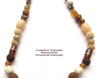 May include: A handcrafted necklace featuring a variety of beads in shades of brown, cream, and amber. The necklace includes round, oval, and textured beads, with a large cream-colored central bead. The text "Création Sylvette Raisonnier Artisanat français" is visible.