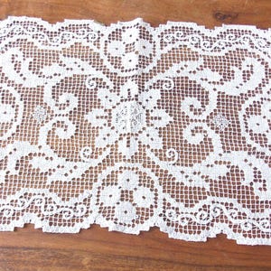Puede incluir: Un tapete rectangular de encaje blanco con un intrincado diseño floral y de volutas. El delicado patrón calado es visible en toda la pieza, con un borde festoneado. El tapete está colocado sobre una superficie de madera.